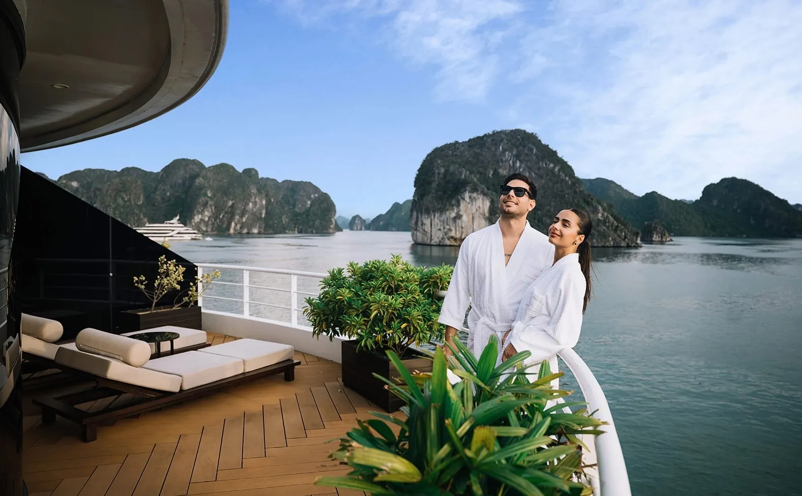 A couple in white robes stands at the railing of a luxury cruise ship deck with sun loungers behind them and karst limestone islands rising from calm water in the background.