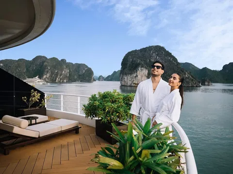 A couple in white robes stands at the railing of a luxury cruise ship deck with sun loungers behind them and karst limestone islands rising from calm water in the background.