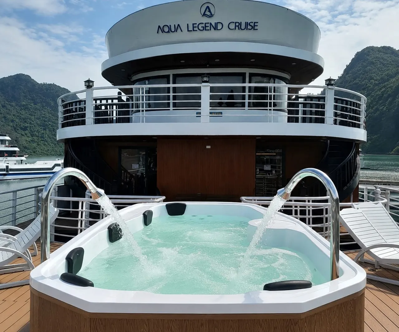 A jacuzzi with two water spouts on a wooden sundeck, with sun loungers and the upper deck of a boat bearing the 'Aqua Legend Cruise' name and logo, set against green karst hills and a Vietnamese flag.