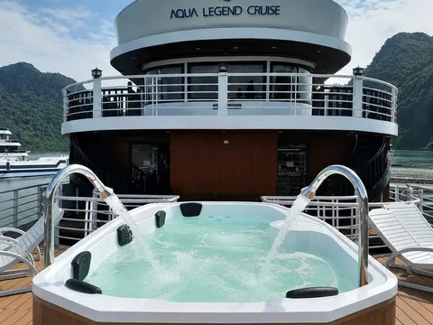 A jacuzzi with two water spouts on a wooden sundeck, with sun loungers and the upper deck of a boat bearing the 'Aqua Legend Cruise' name and logo, set against green karst hills and a Vietnamese flag.