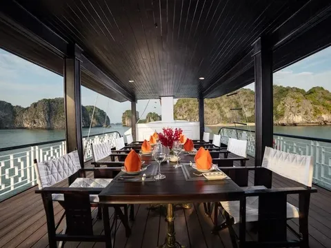 A covered upper deck with a dark wooden dining table set with orange napkins, glasses and red flowers, white railings on both sides, and limestone karst islands across calm water in the background.