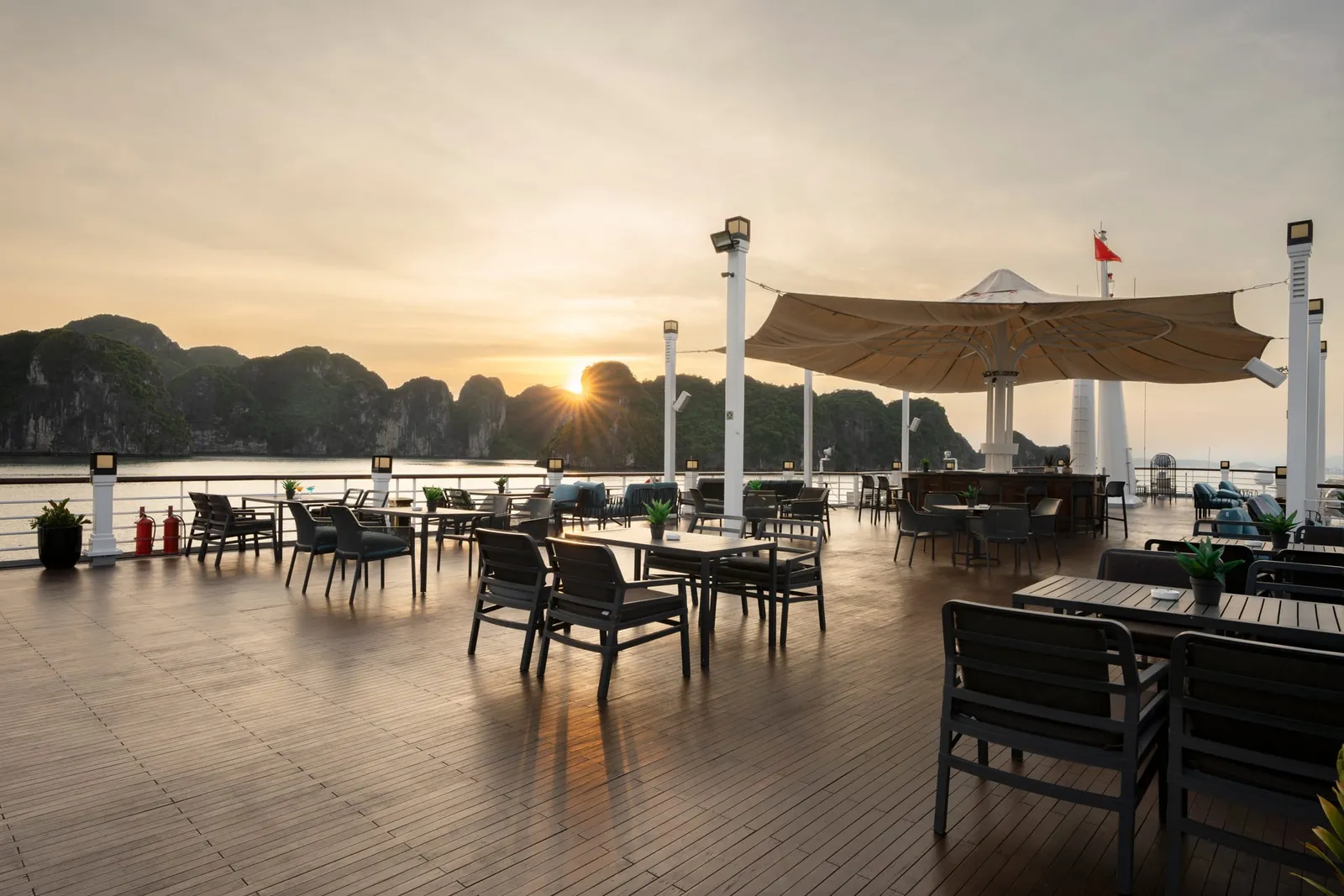 A wooden deck with dining tables and chairs under umbrellas at sunset, with limestone karst islands visible across the water in the background.