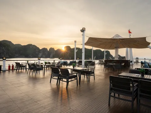 A wooden deck with dining tables and chairs under umbrellas at sunset, with limestone karst islands visible across the water in the background.