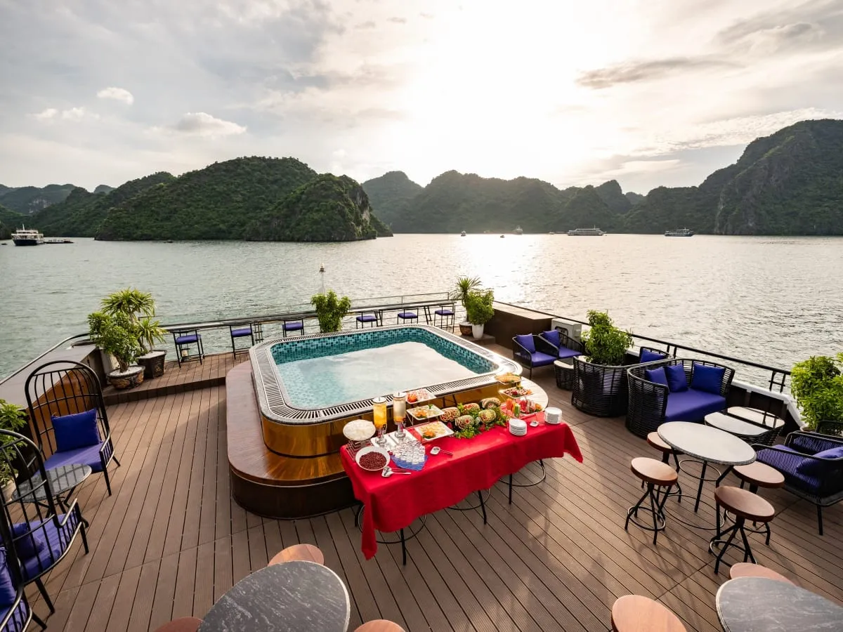 A wooden sun deck with a tiled jacuzzi, a buffet table with a red cloth, blue-cushioned chairs and stools, overlooking calm water and forested limestone islands with distant boats.