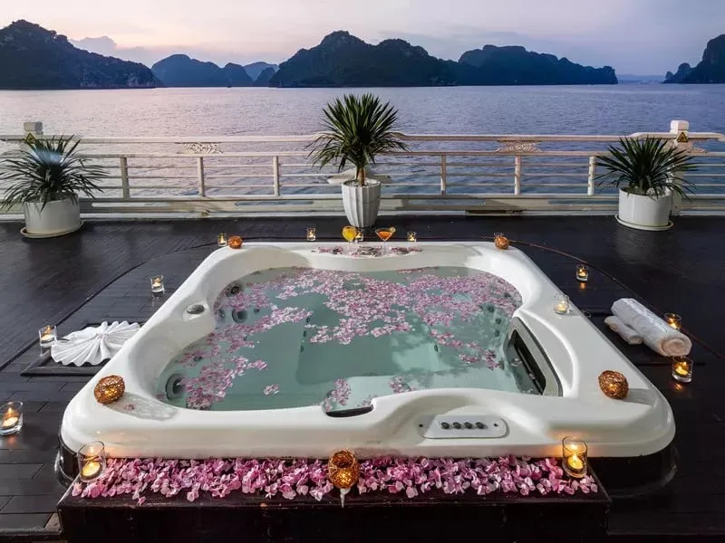 A white hot tub filled with pink flower petals sits on a wooden deck with limestone karsts and calm water in the background at dusk.