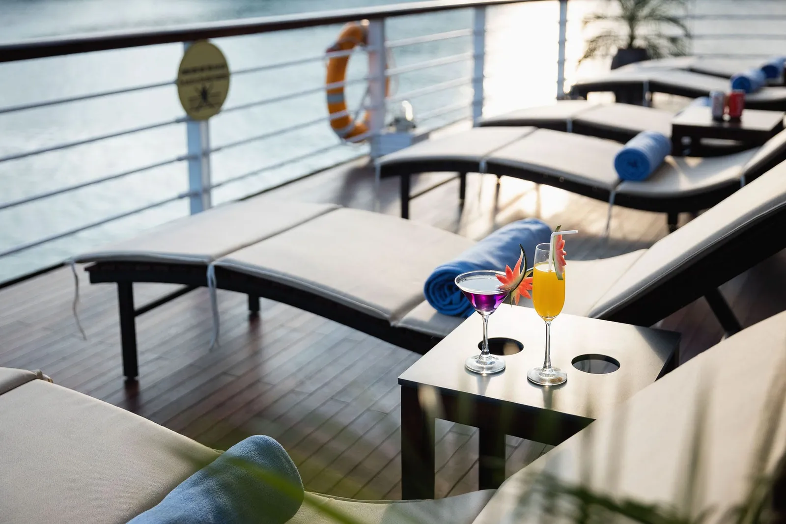 Sundeck with lounge chairs, a small table holding two cocktails, life ring and clock mounted on the railing, and water visible in the background.