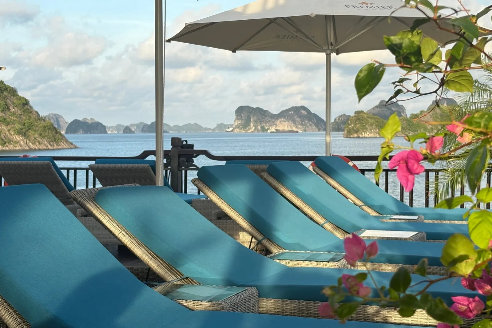 Teal sun loungers under a white umbrella, bougainvillea in the foreground and karst on the horizon