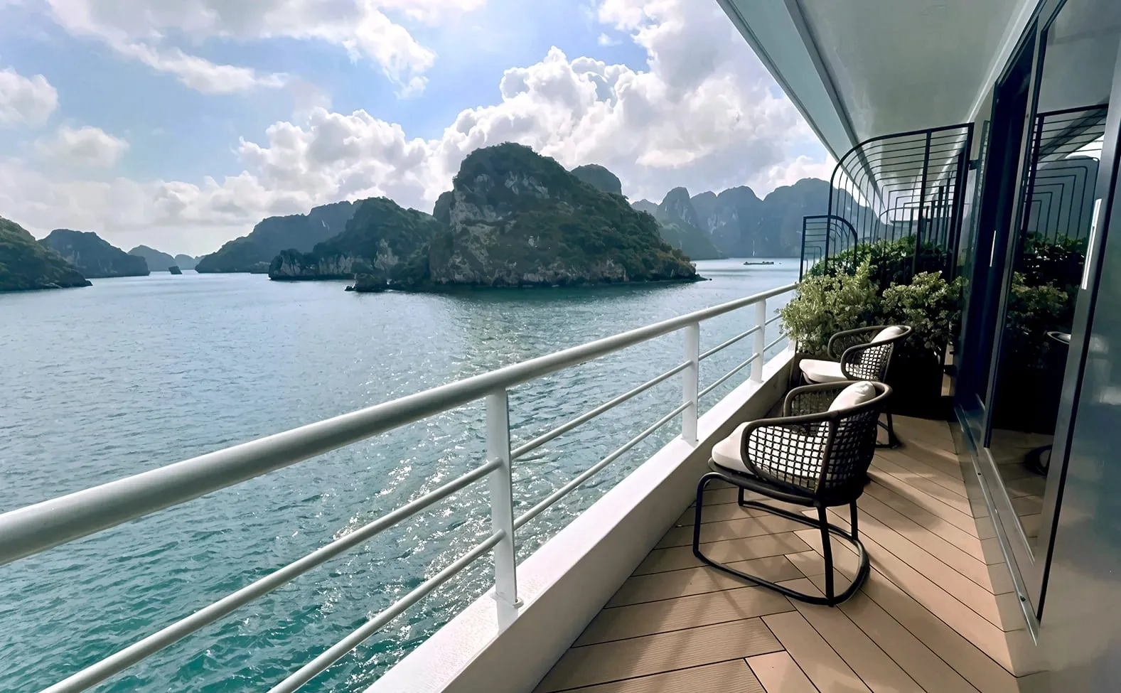 A modern cruise ship balcony with wicker chairs and a wood-plank deck overlooking karst limestone islands rising from calm turquoise water.
