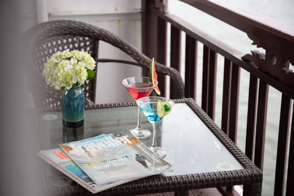 A wicker chair and glass-topped wicker table on a wooden-railed balcony holds two cocktails, a vase of white hydrangeas and travel magazines, with calm water visible beyond the railing.