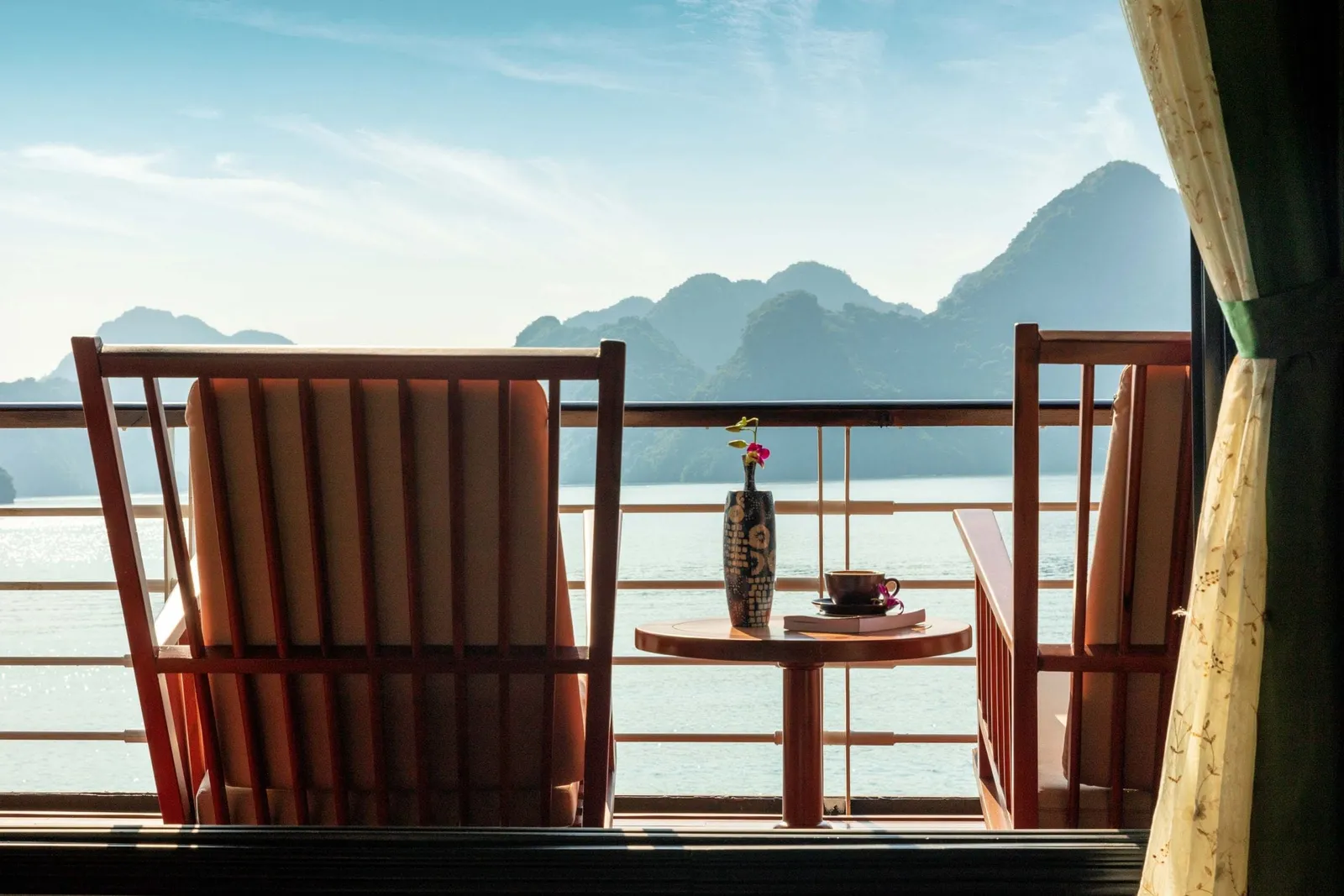 Two wooden chairs with cushions and a small round table holding a vase with a flower, a cup and a book on a balcony railing, facing calm water and misty limestone karst peaks, with a curtain at the right edge.