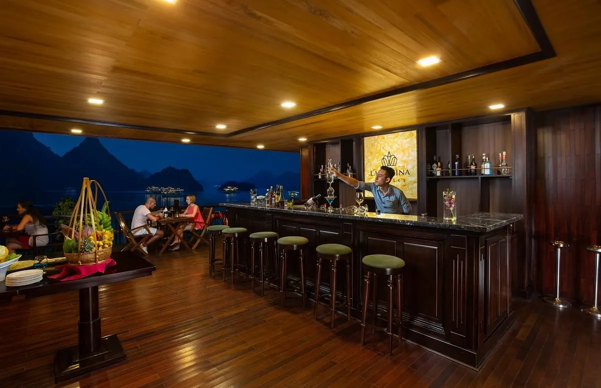 An indoor bar area on a cruise boat with wooden floors and ceiling, green bar stools, a bartender in blue, and an open deck area behind showing limestone karsts at dusk.