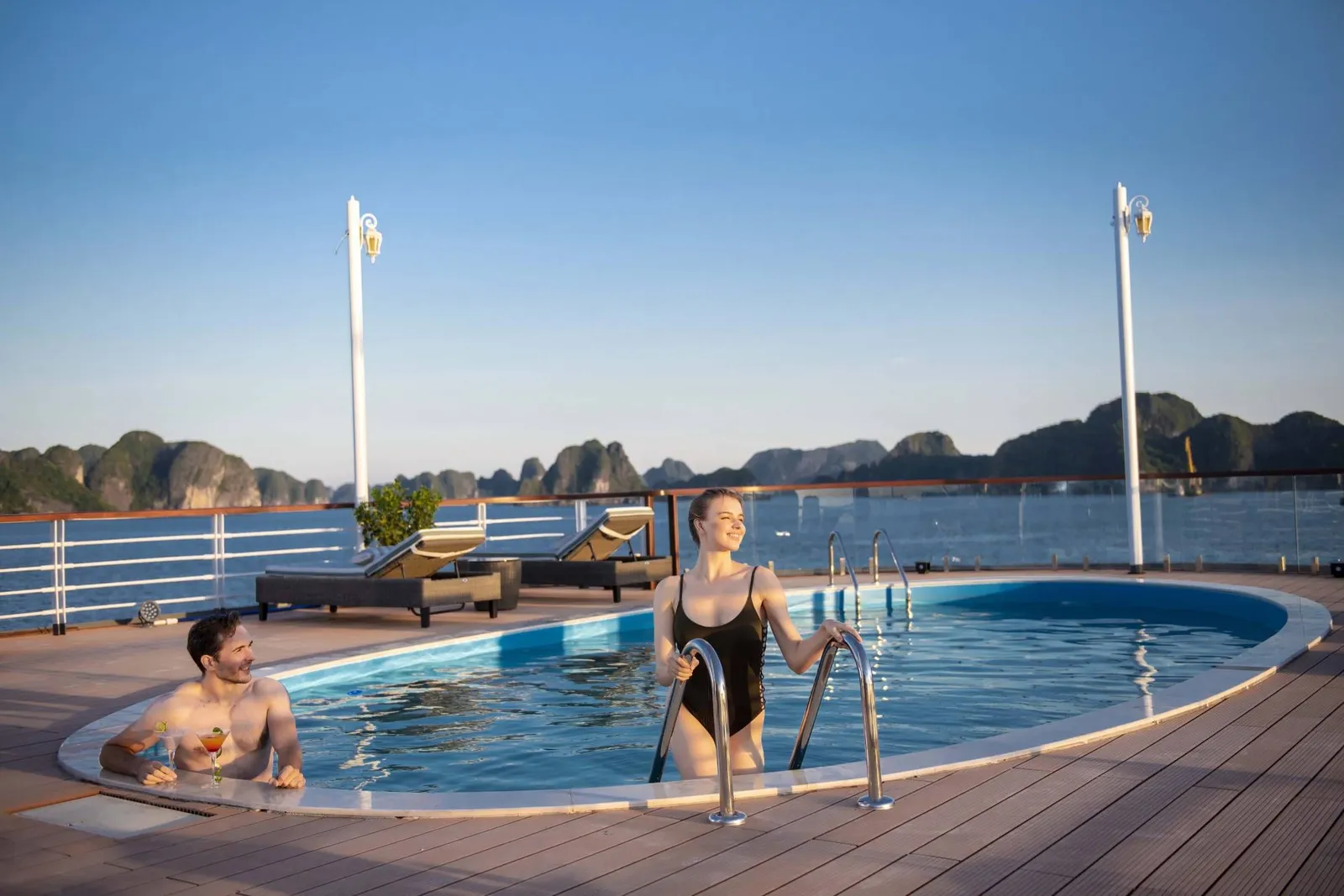 Two people in a swimming pool on a boat deck with limestone karsts visible across the water in the background.