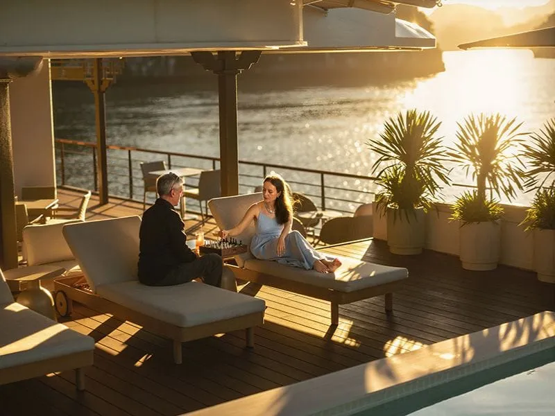 Two people sit on lounge chairs on a wooden deck with a small pool, potted palms, and sparkling water visible beyond the railing at golden hour.