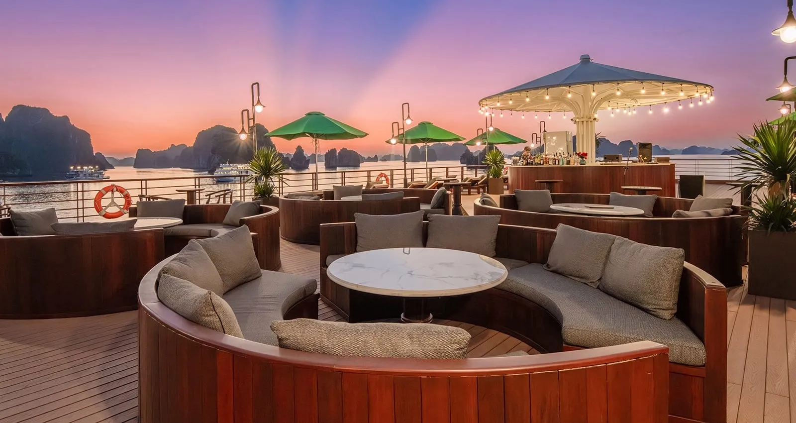Sundowner rooftop deck with curved wooden banquette seating, marble tables, green umbrellas and a bar under a lit canopy, with limestone karsts and distant boats at dusk.