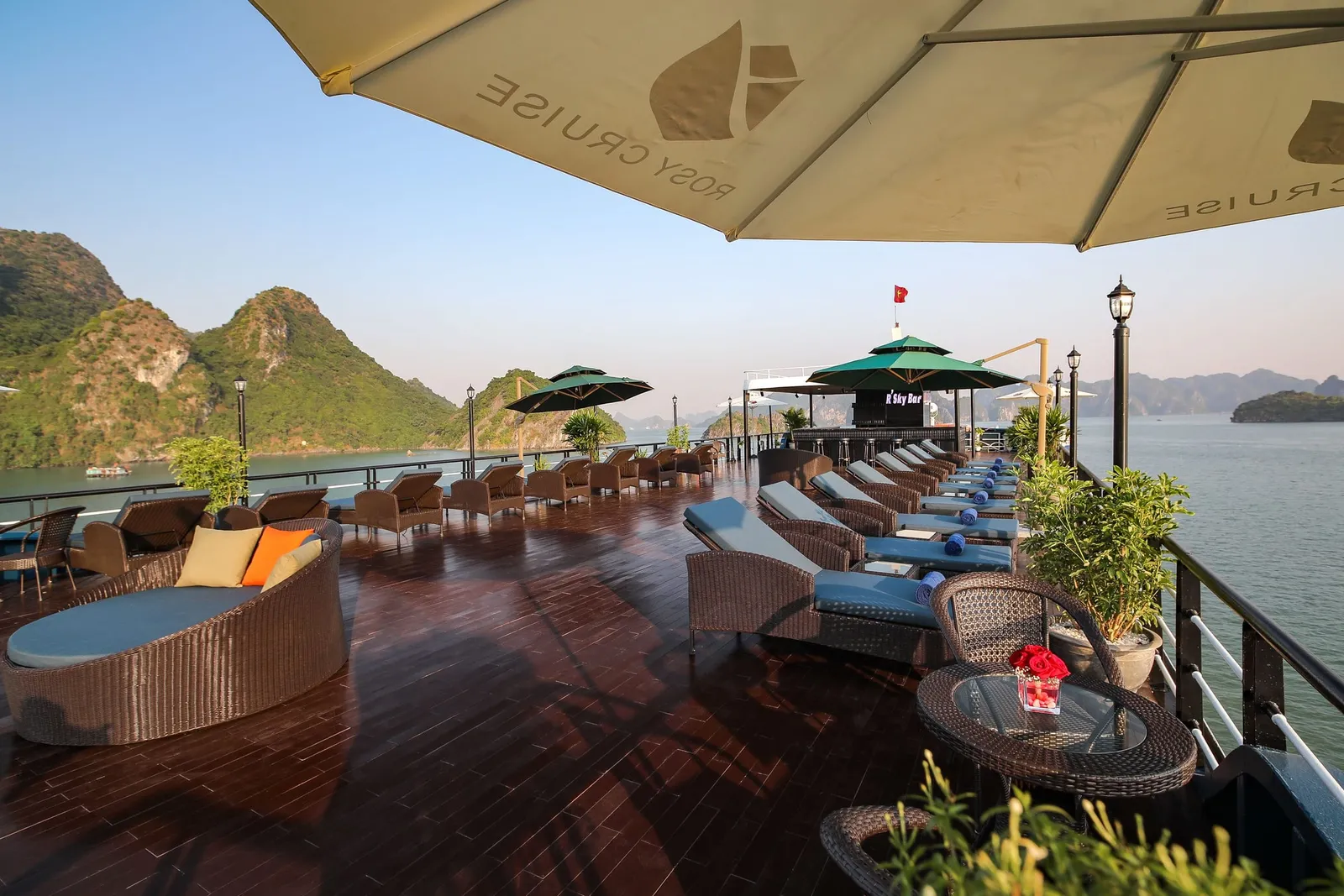 Sundeck lined with loungers under branded parasols, a low table with a red flower in front, the bay and karst beyond the rail