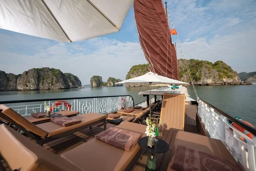 Sun deck of a junk-style boat with cushioned loungers, folded towels embroidered with a logo and text, white umbrellas, a brown sail and limestone karst islands across calm water.