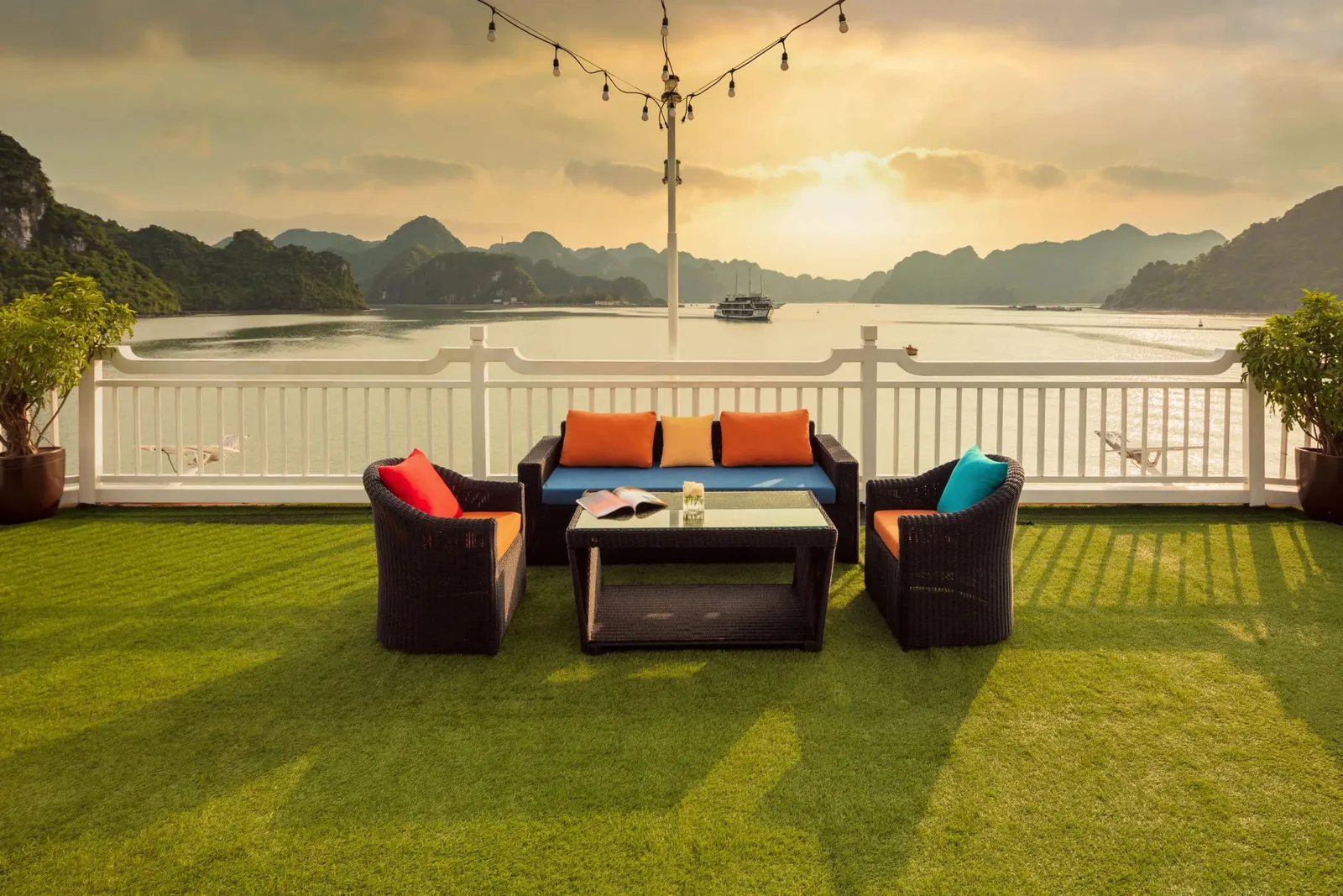 A sun deck with artificial grass, dark wicker sofa and two armchairs with orange and teal cushions, a white railing, and a bay with limestone karst islands and a distant boat at sunset.