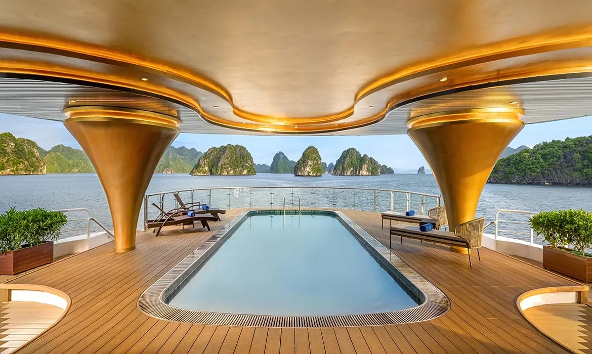 A luxury cruise ship's upper deck features a rectangular pool, teak decking, lounge chairs, and gold-toned structural columns under a curved canopy, with karst limestone islands visible across the water in the background.