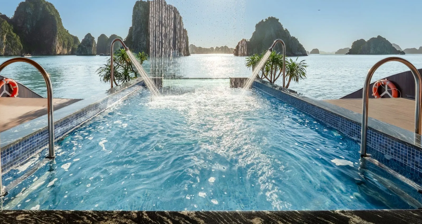 A blue-tiled pool on a ship's upper deck with water jets and steel handrails, life rings on the sides, and limestone karsts rising from the bay in the background.