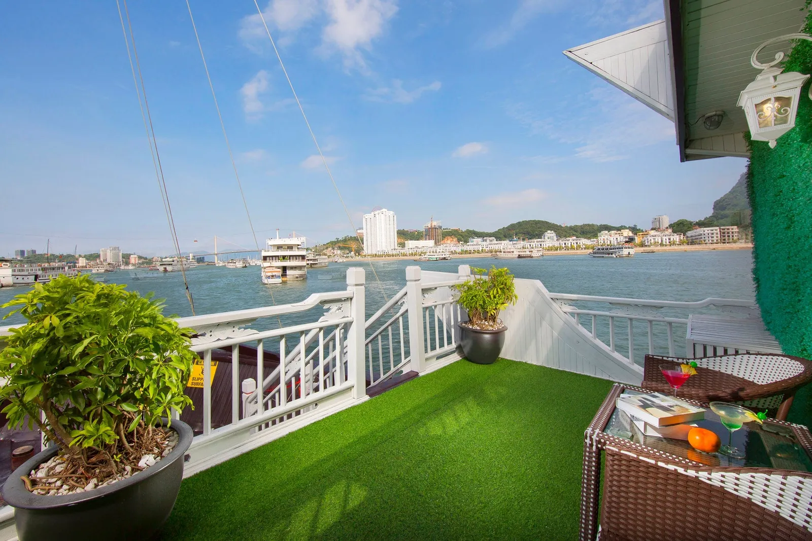 A small balcony deck with artificial grass, white railings, potted plants and wicker chairs with cocktails on a table, overlooking a harbour with boats, a bridge and waterfront buildings.
