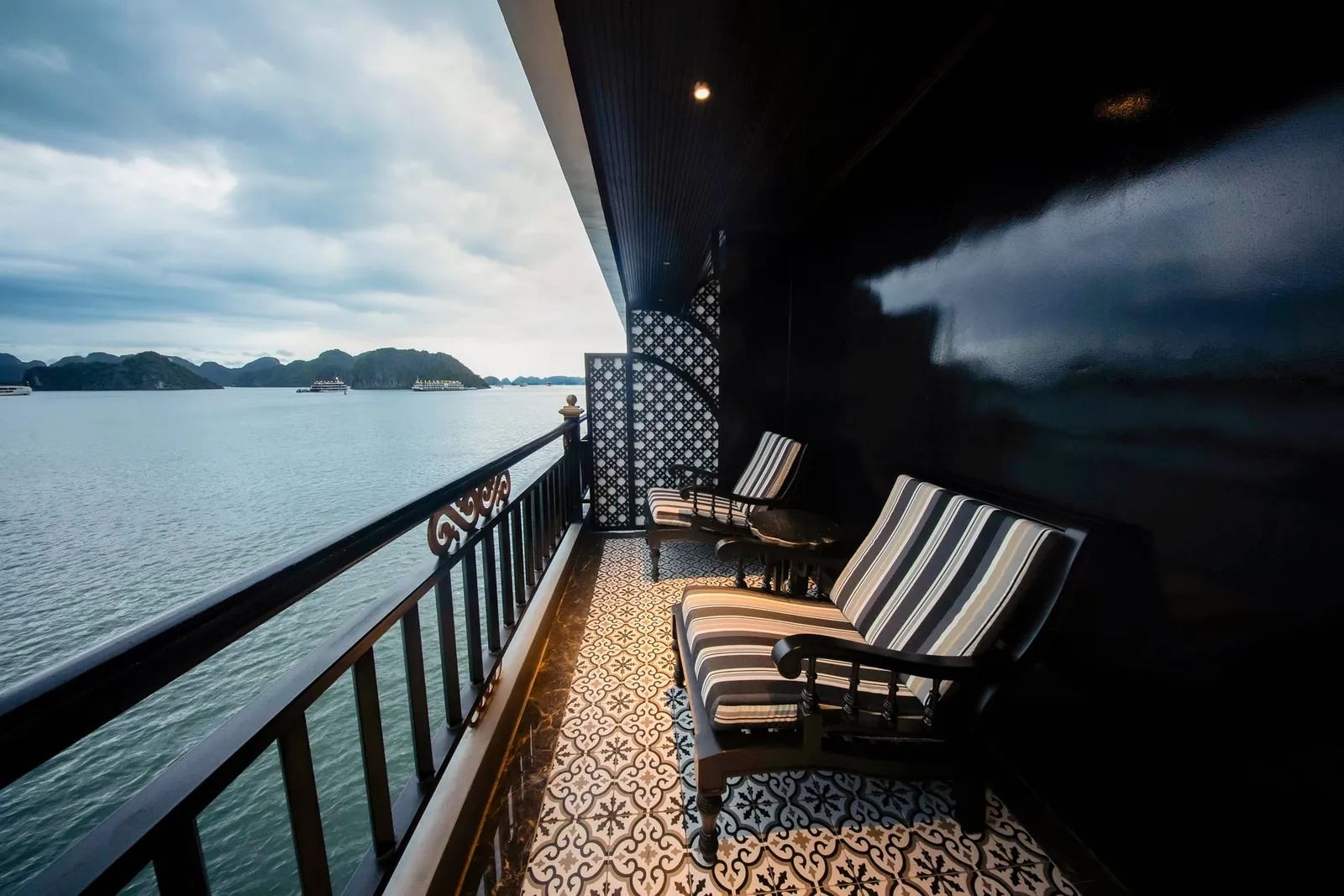 A private balcony on a boat with two striped-cushion chairs, patterned floor tiles and a dark railing, looking out over calm water toward limestone islands and distant boats under a cloudy sky.