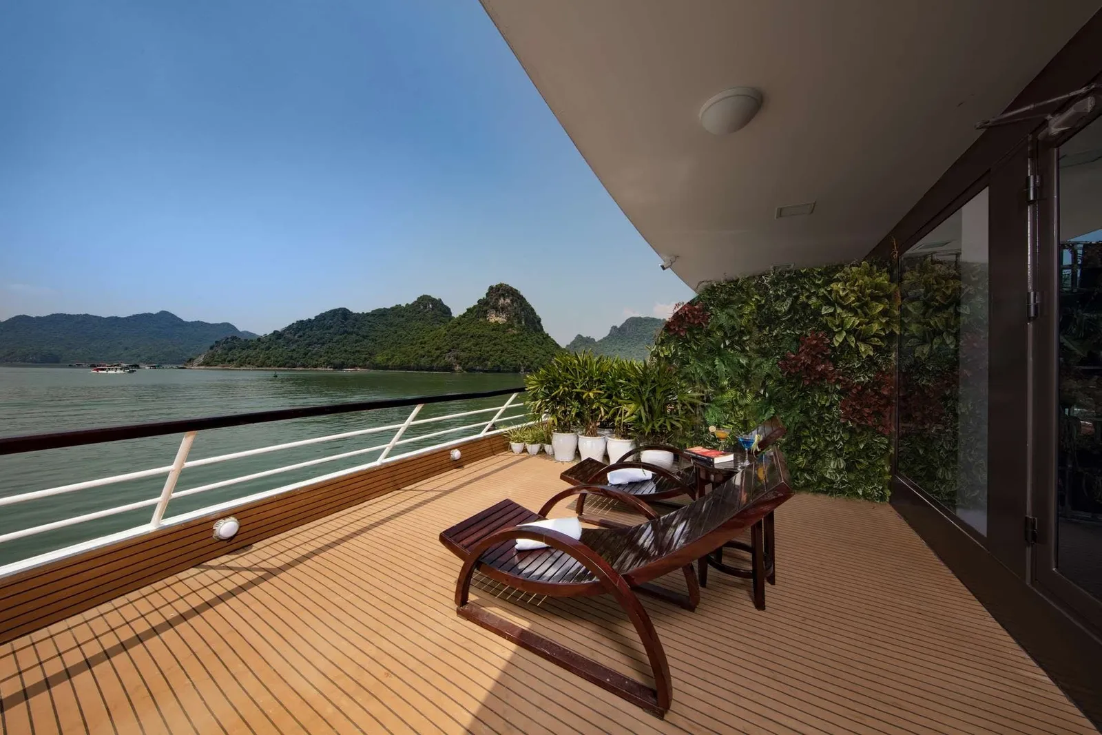 A teak private balcony deck with two wooden lounge chairs, potted plants and a green living wall, overlooking calm green water and forested limestone karst hills.