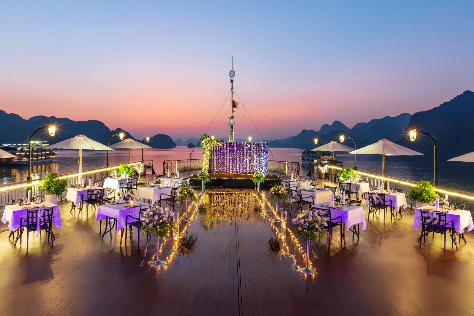 Top deck laid out for dinner at sunset, tables under purple cloths and lights strung along the rail