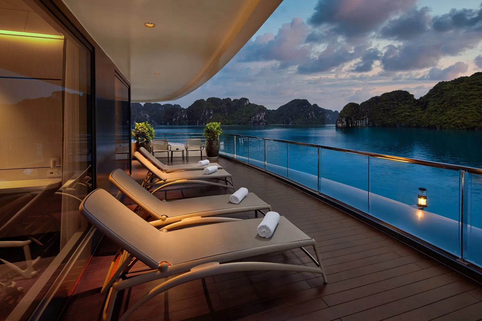 A private balcony deck of a cruise ship at dusk with four lounge chairs with rolled towels, glass railing, a lantern, and limestone karst islands across calm blue water.