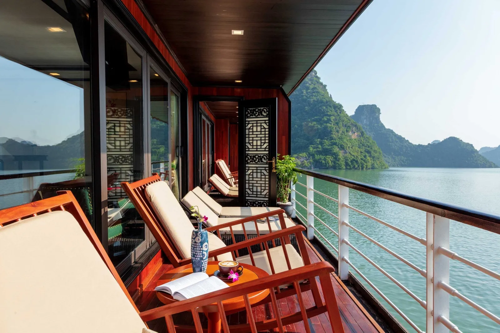 A wooden side deck of a cruise ship with cushioned loungers, a small table with coffee and an open book, and a white railing overlooking green water and forested limestone karst islands.