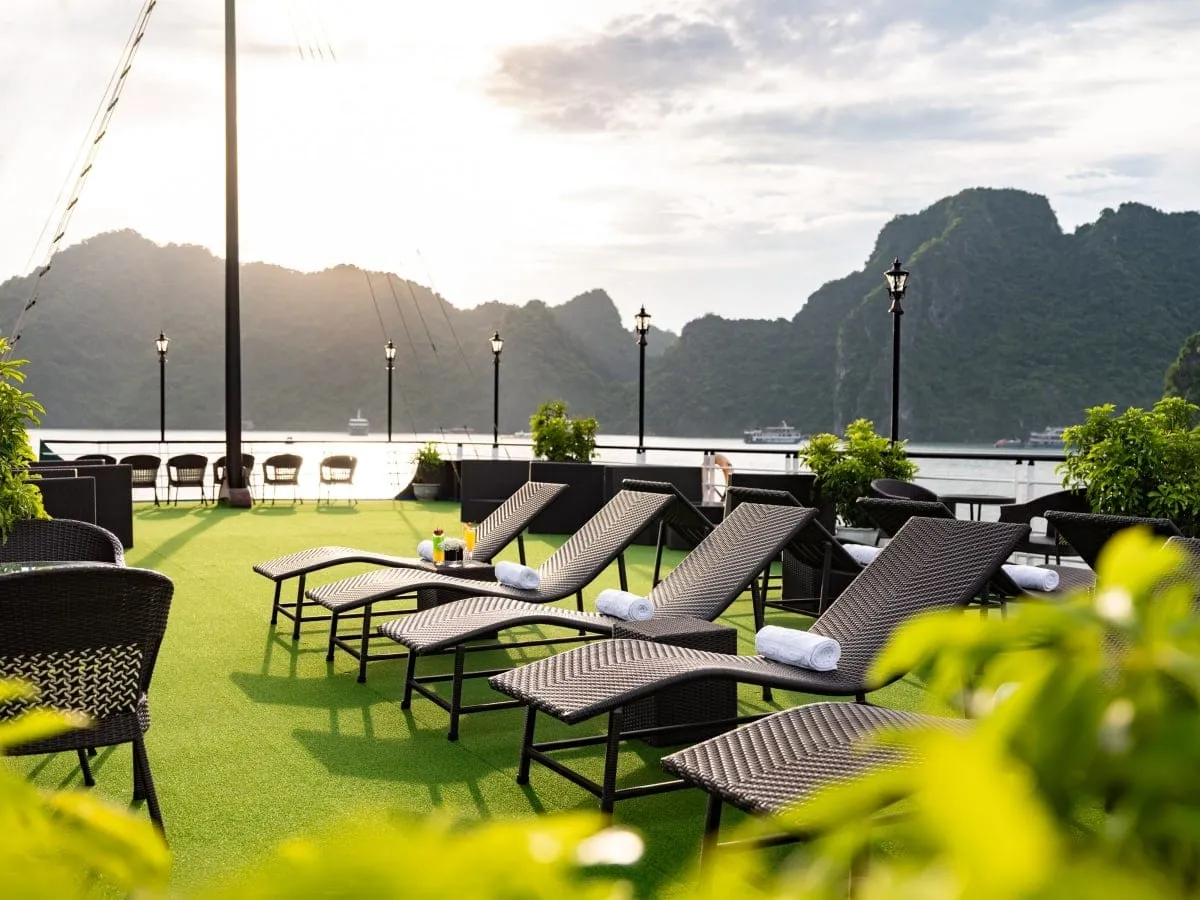 Sun deck with artificial grass, wicker loungers with rolled towels and drinks, lamp posts and potted plants, with limestone karst hills and distant boats across the bay.