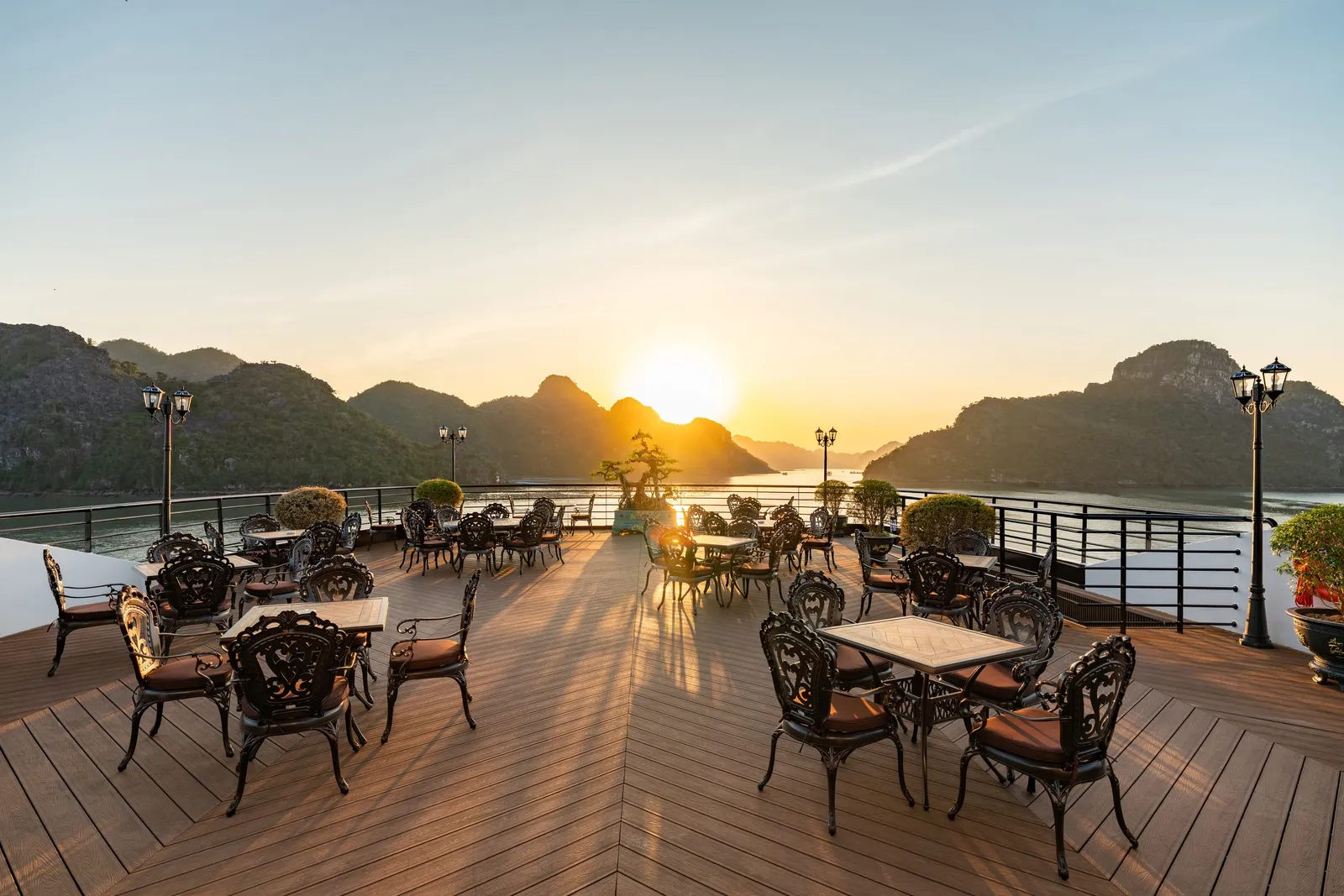 A wooden deck with ornate dark chairs and tables arranged for dining faces dramatic limestone karst islands at sunset.