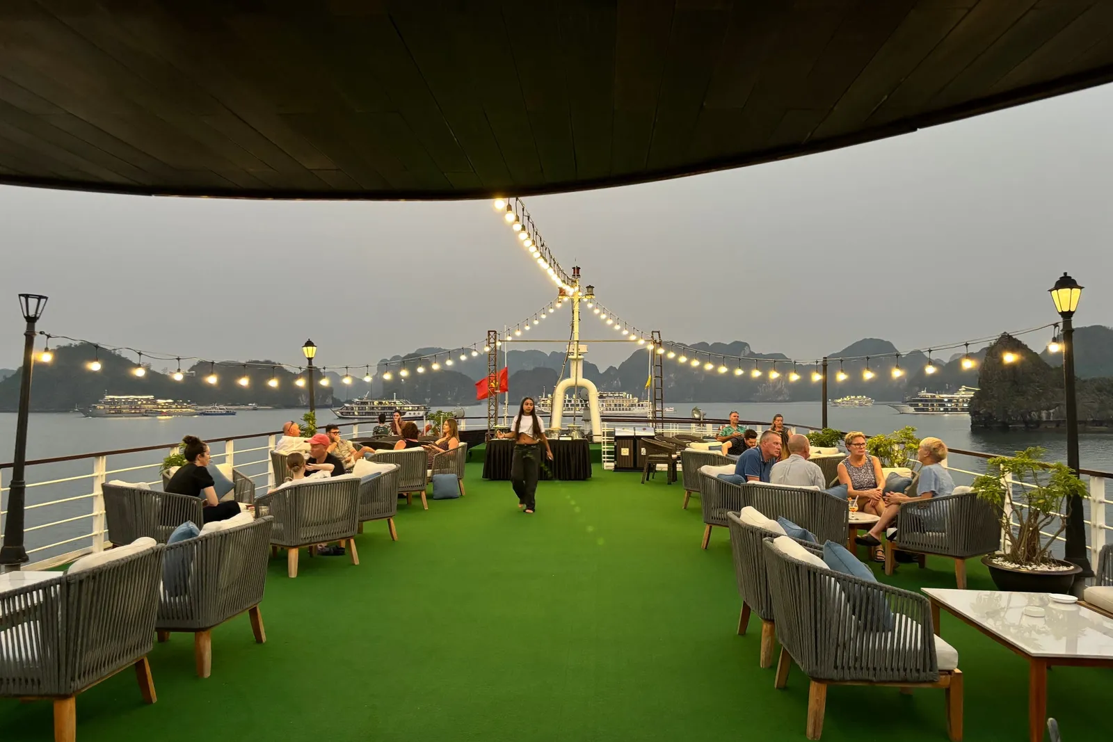 Upper deck at dusk laid with green turf, armchairs in groups and lights strung the length of the ship
