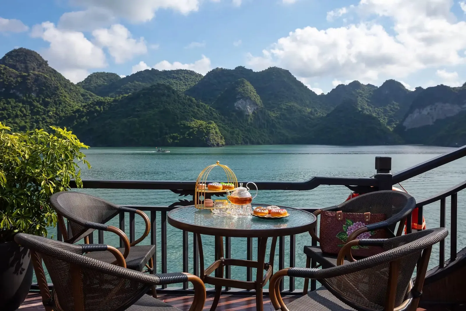A glass-top table with a teapot, glasses and a tiered pastry stand sits on a wooden deck with rattan chairs and a dark railing, overlooking a green bay with forested limestone hills and a small distant boat.