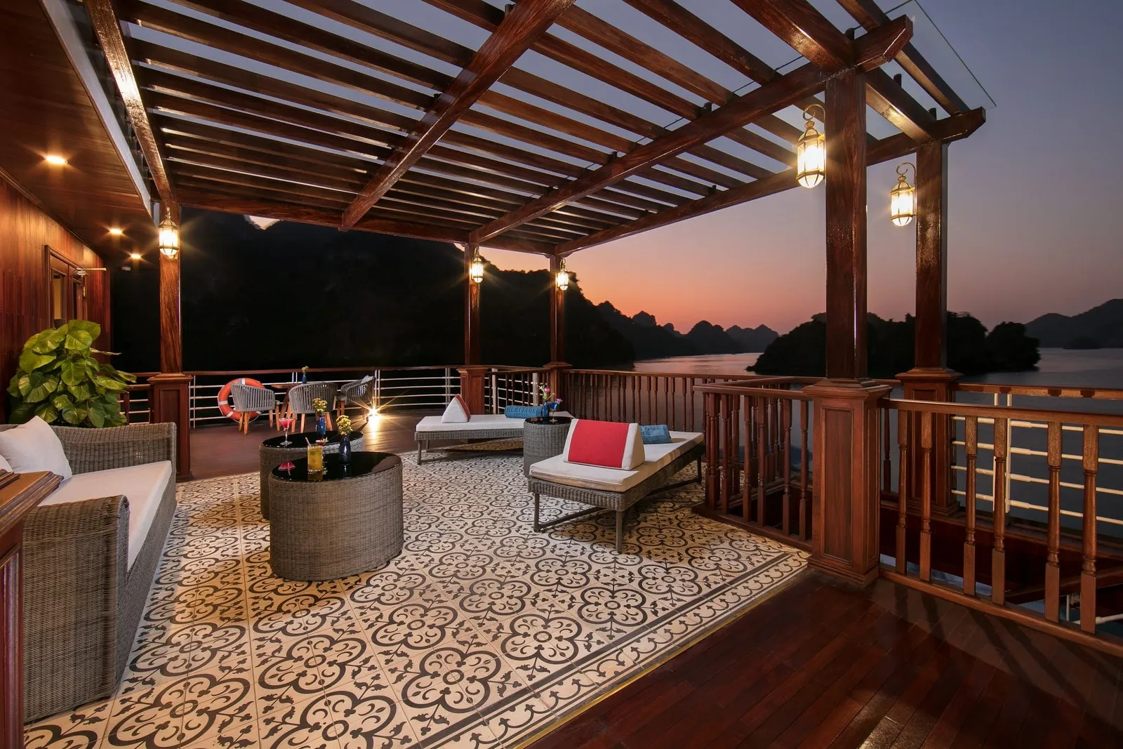 A covered wooden upper deck with a pergola, wicker lounge furniture, patterned tile floor and lanterns at dusk, overlooking karst islands in calm water.