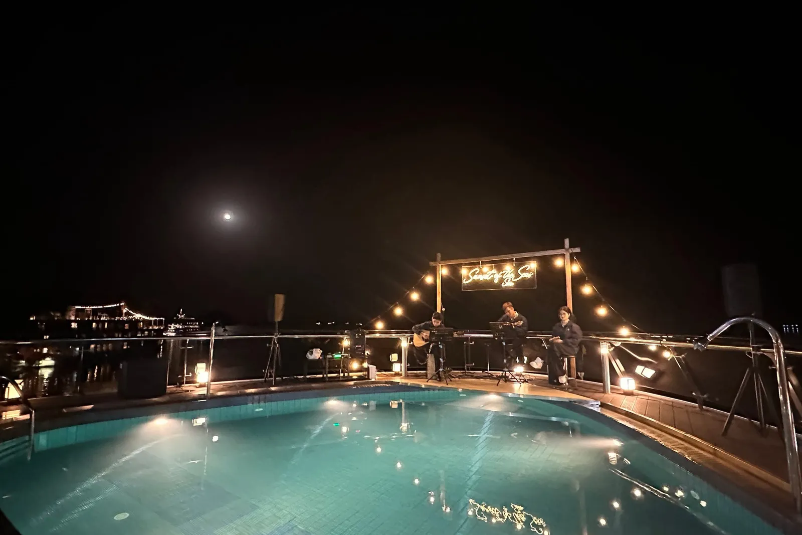 Pool deck after dark under strings of lights, guests standing at the far rail
