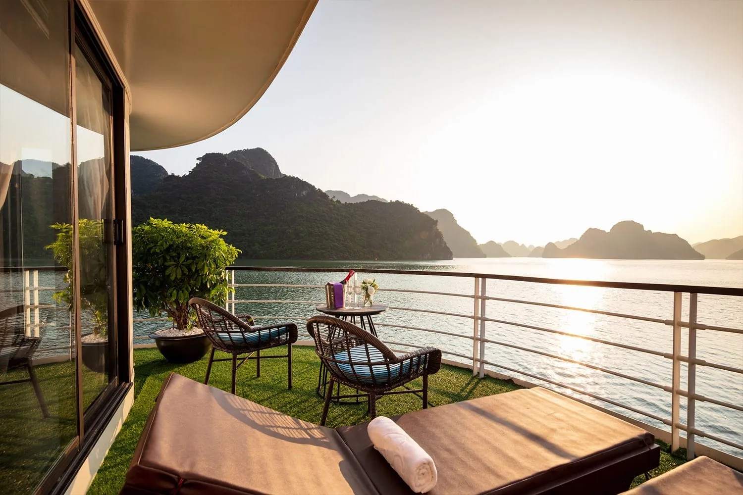 A private balcony terrace with artificial grass, two rattan chairs, a small table with a champagne bucket and glasses, and a sun lounger, facing limestone karsts in a sunlit bay.