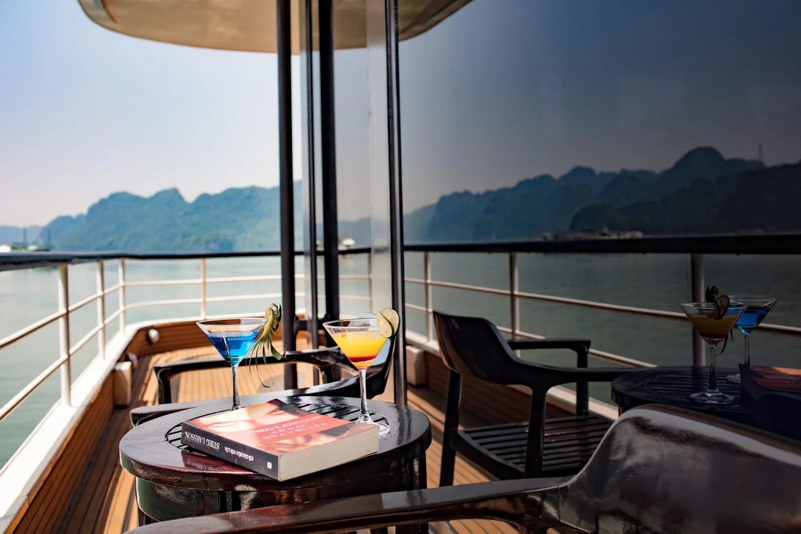 A wooden ship deck with small tables, chairs, cocktails and a paperback book, overlooking calm water and forested limestone hills.