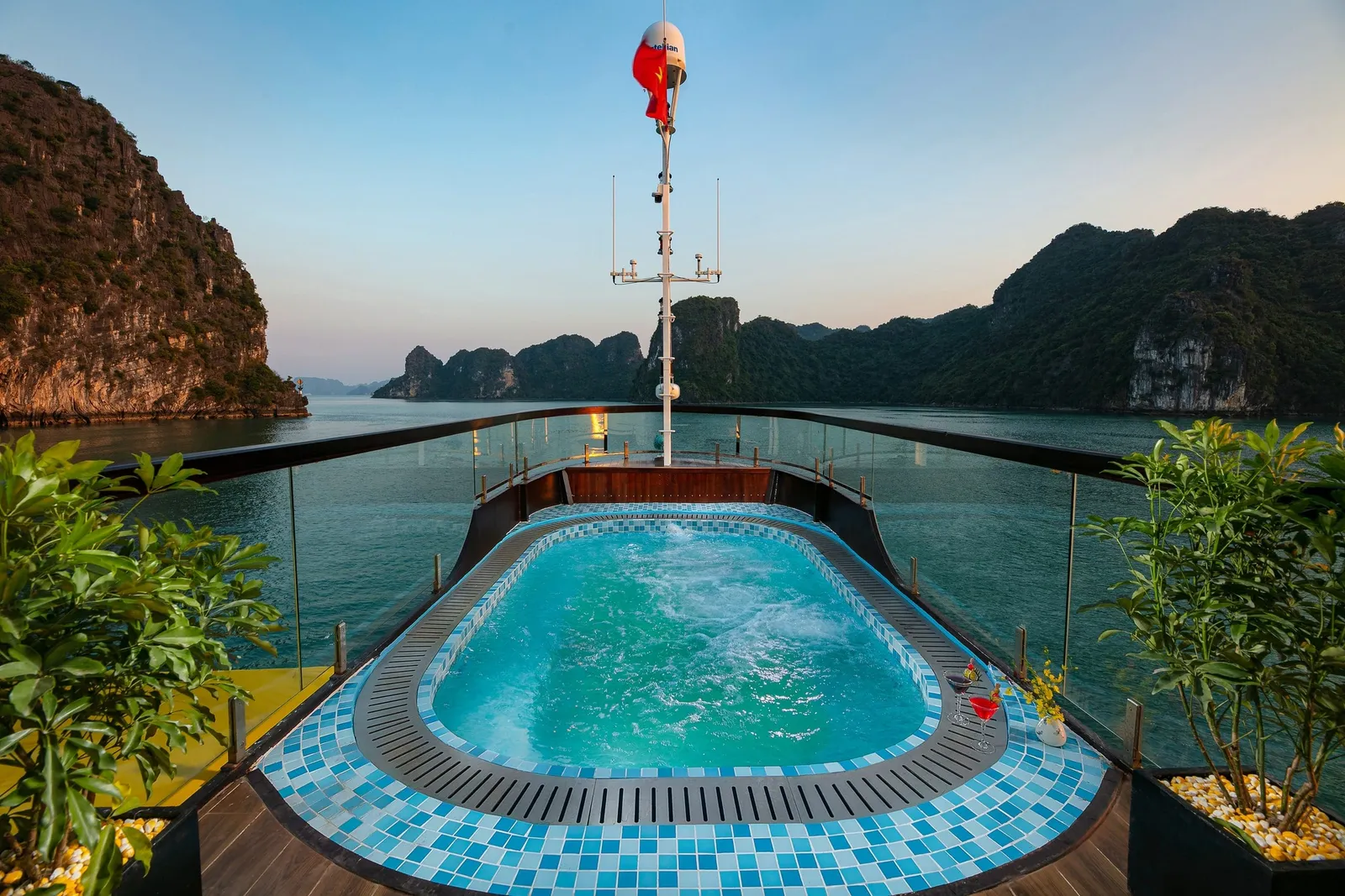 A tiled jacuzzi pool on the upper sun deck of a ship with glass railings, a mast with a Vietnamese flag, and limestone karst islands across calm water at dusk.