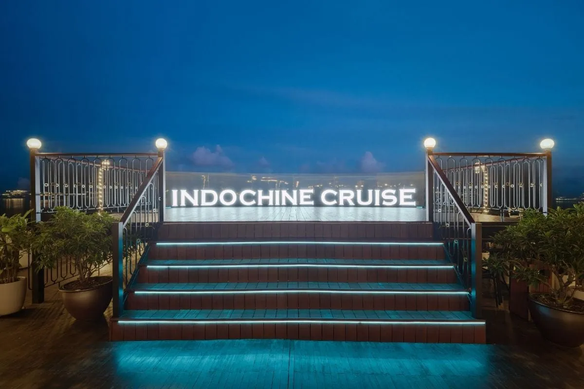 A dusk view of a ship's upper deck with an illuminated staircase leading to a platform with a lit 'INDOCHINE CRUISE' sign, flanked by railings, lamps and potted plants.
