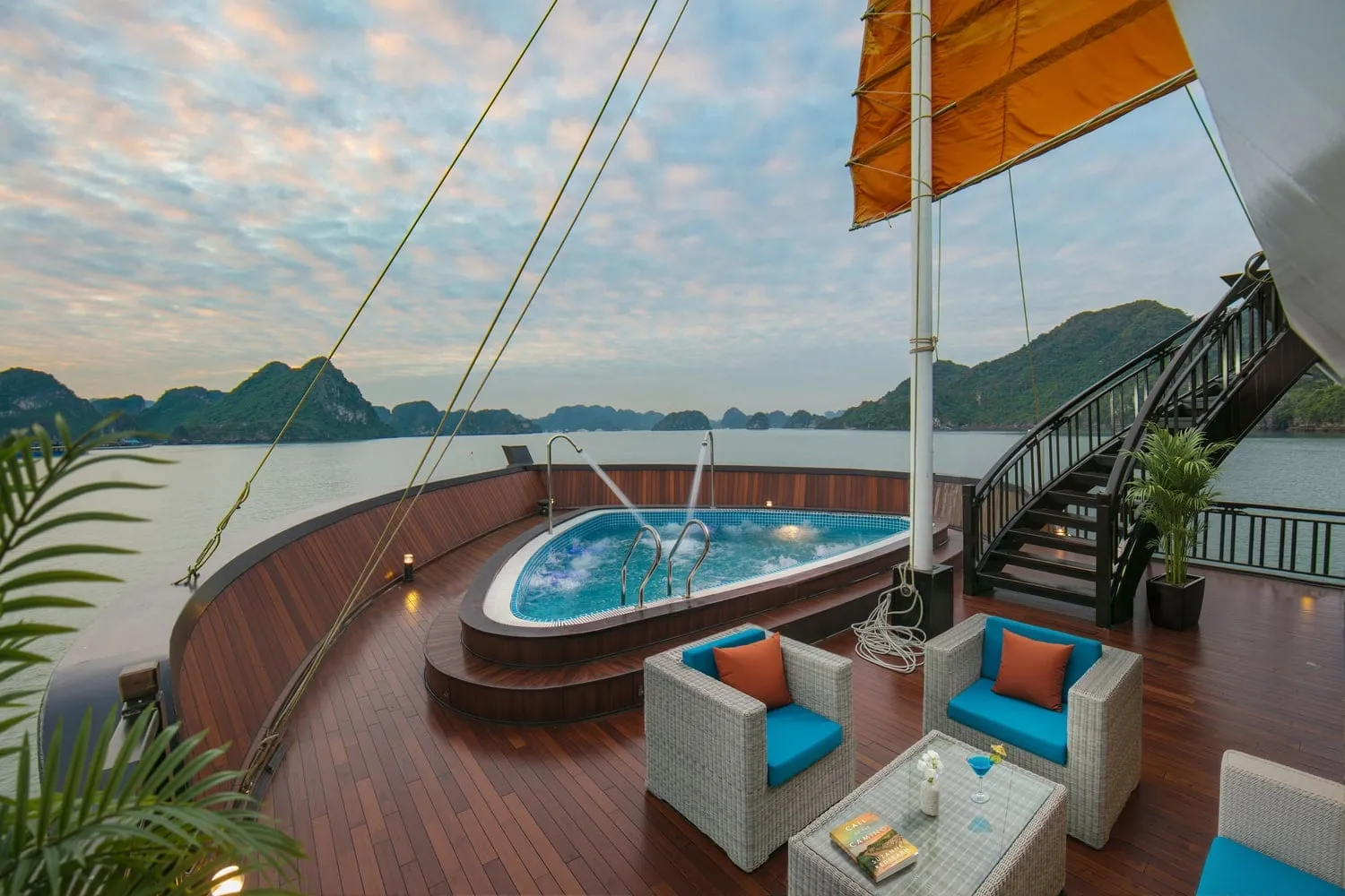 Wooden bow deck of a junk-style ship with a jacuzzi pool, wicker chairs with teal and orange cushions, an orange sail and a staircase, overlooking limestone karsts at dusk.