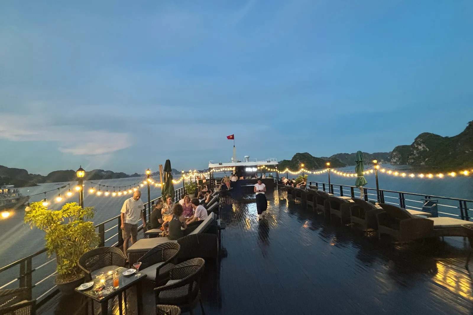 Deck at dusk under strings of lights, guests at the tables and the pool lit beside them