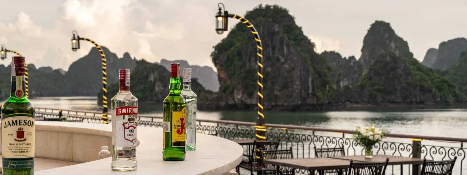 Bar counter on a ship's deck with Jameson, Smirnoff and other liquor bottles, with tables, chairs and yellow-black striped lamp posts overlooking karst limestone islands in a bay.