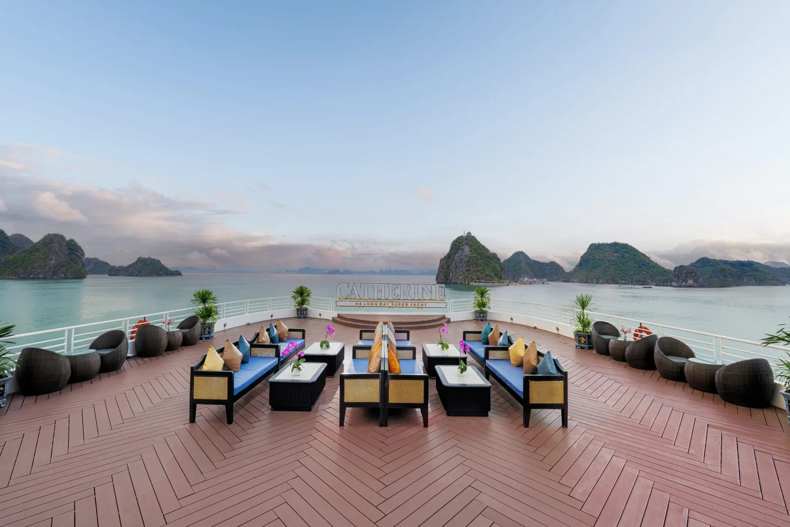 Sun deck with a herringbone wooden floor, blue-cushioned sofas, wicker chairs and potted plants, with a 'CATHERINE Ha Long Bay Super Yacht' sign at the rear railing and limestone karsts across the water.