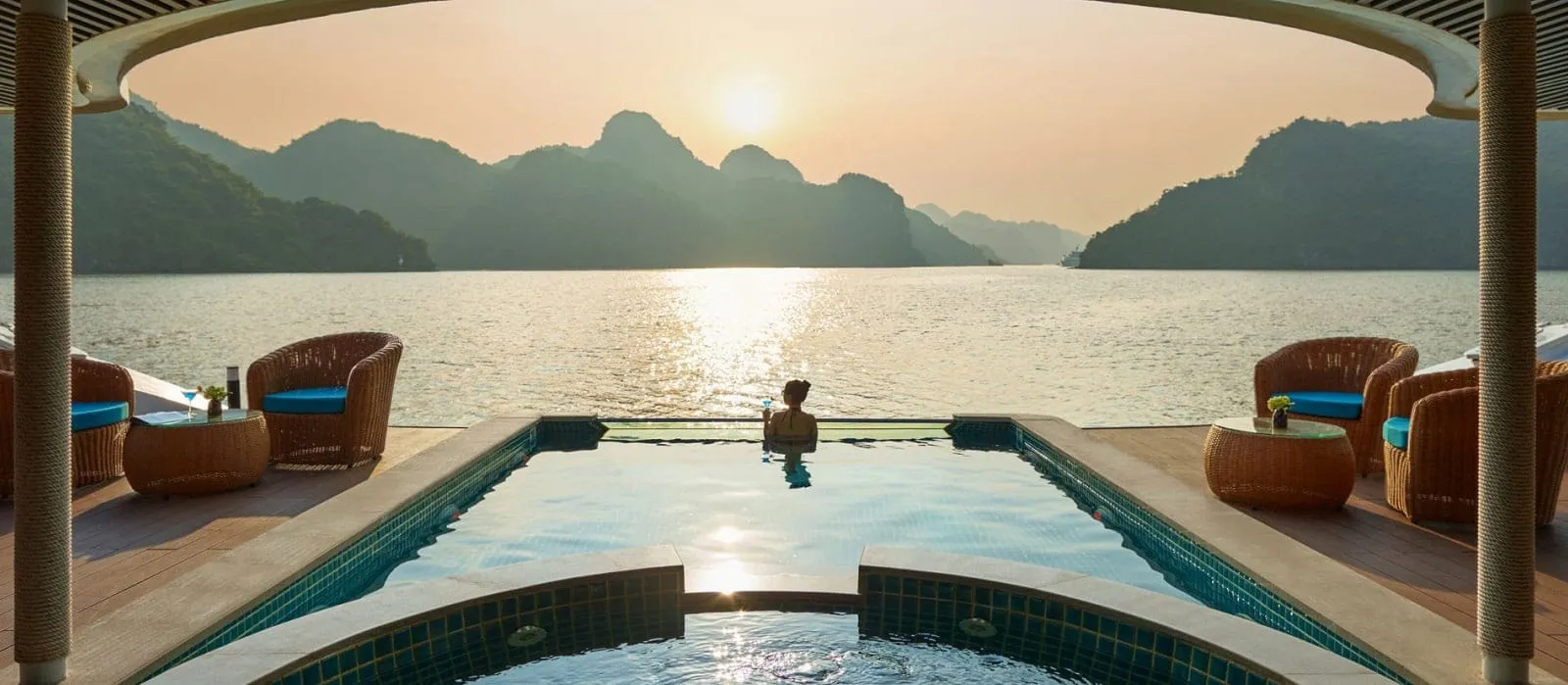 A person stands in an infinity pool on a ship's upper deck with wicker chairs on either side, facing limestone karst islands and the sun setting over the water.