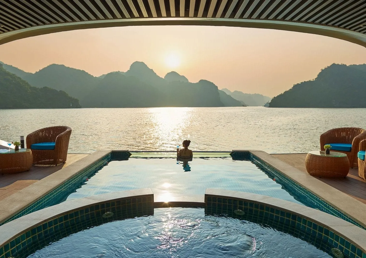 A pool deck with a round jacuzzi and infinity pool under a slatted canopy, with a person in the water facing a sunset over limestone karst islands, flanked by wicker chairs and side tables.