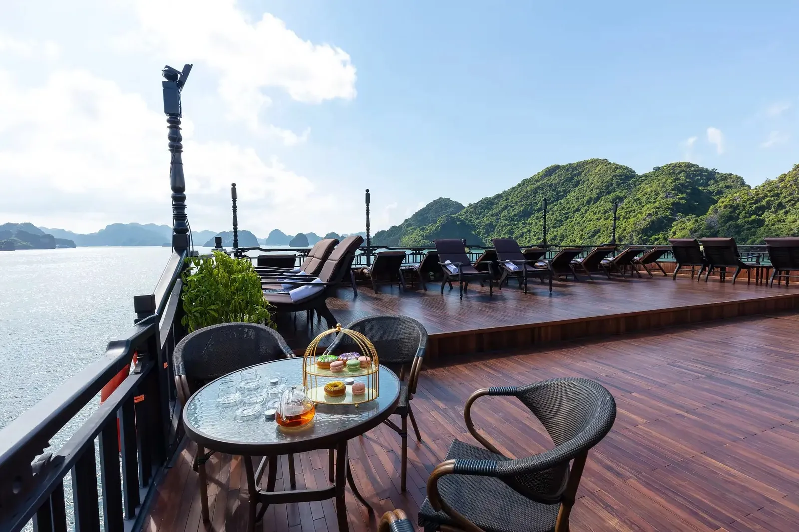 A wooden sundeck with a glass table holding tea and a donut stand, wicker chairs, rows of sun loungers, and a railing, with green karst islands and calm water beyond.