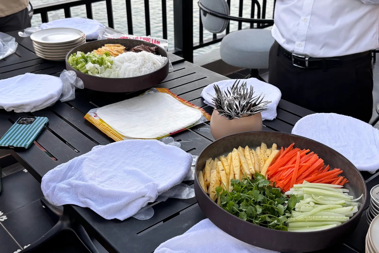 A deck table set up for a spring-roll making session with bowls of sliced pineapple, carrot, cucumber, herbs, noodles, rice paper and folded towels, two staff standing by a railing with calm water and a white cruise boat behind.