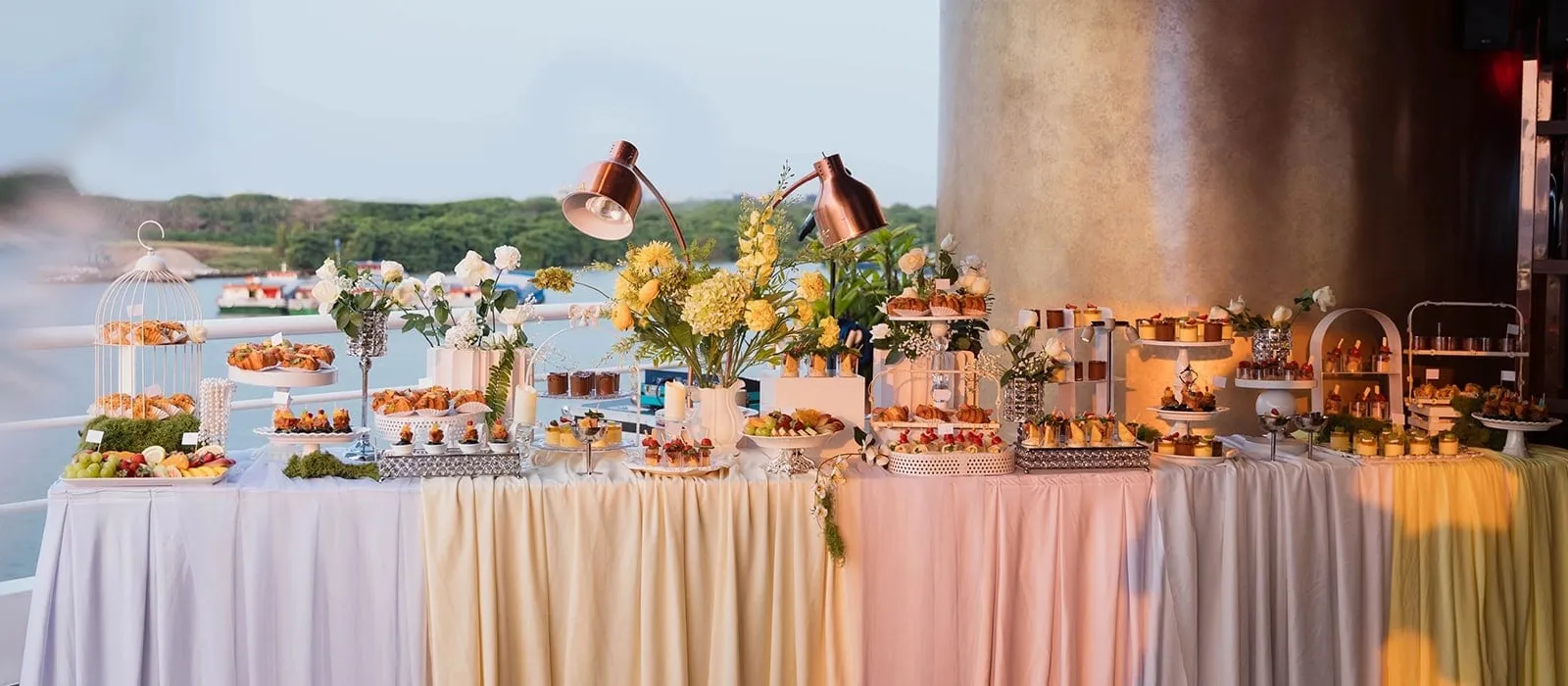 A long buffet table with pastel draped cloths holds tiered dessert and canapé stands, yellow flowers and heat lamps, on a deck with a railing and a green shoreline and small boats behind.