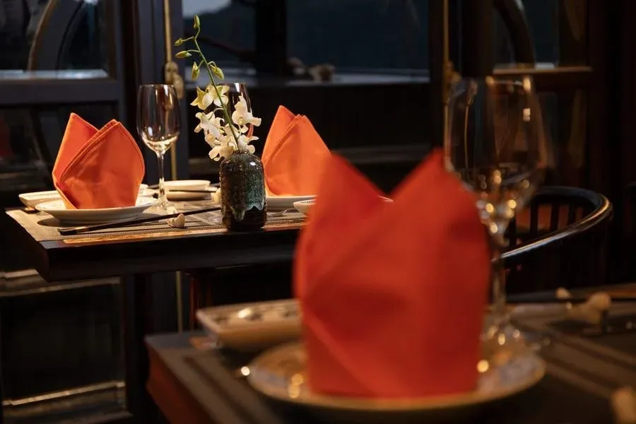 Dark wooden dining tables set with folded orange napkins, wine glasses, plates, chopsticks and a small vase of white orchids, in a dimly lit room with window frames behind.