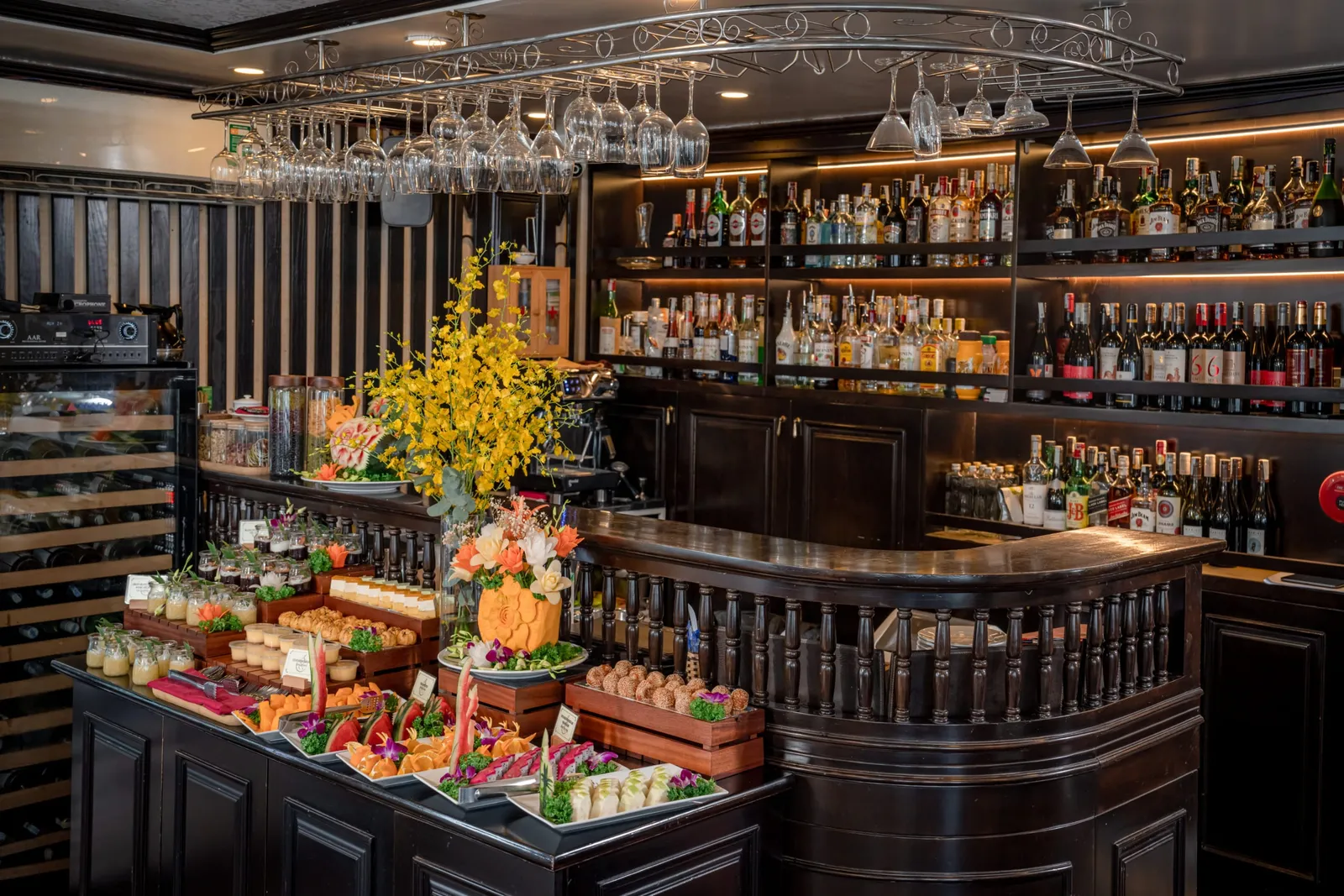 A dark-wood bar with shelves of bottles and hanging wine glasses, and a buffet counter in front displaying small desserts, carved fruit, canapés and flower arrangements with yellow orchids.